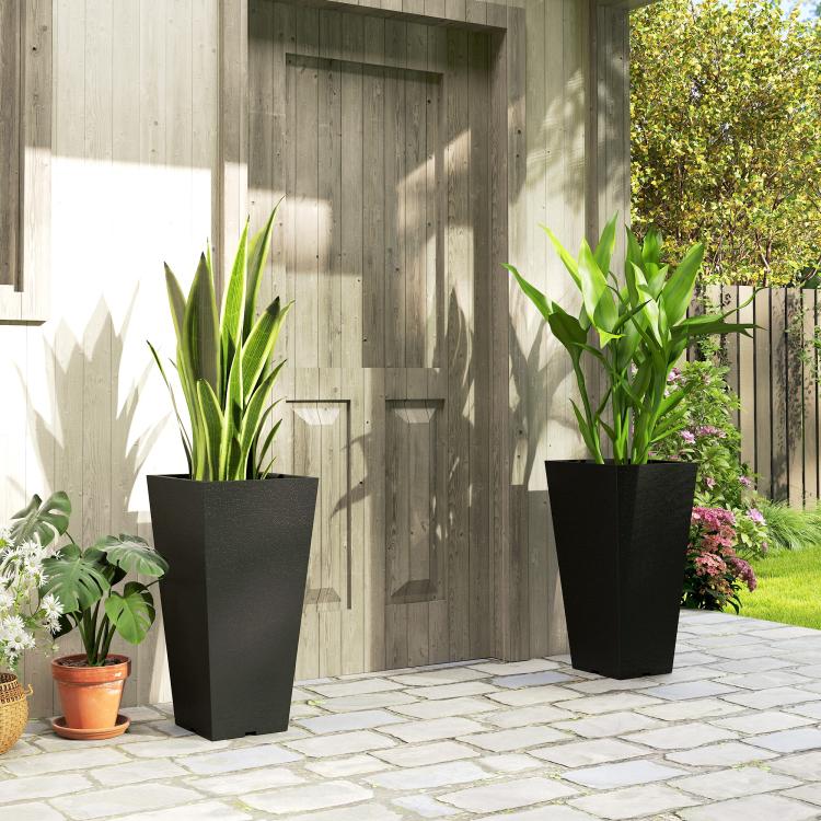 Two tall black square planters with green plants beside a walkway