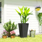 Tall black outdoor planter with green bamboo-like plant on grass near flowers