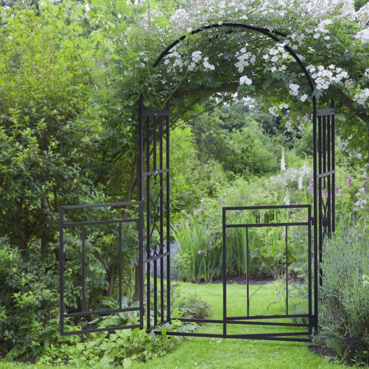 Grey metal garden arbor arch with double doors open, framed by greenery and flowers
