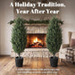 Two faux cedar mini trees in black textured pots in front of a fireplace