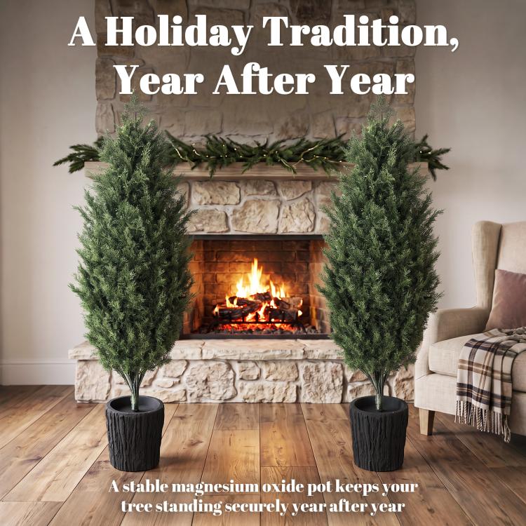 Two faux cedar mini trees in black textured pots in front of a fireplace