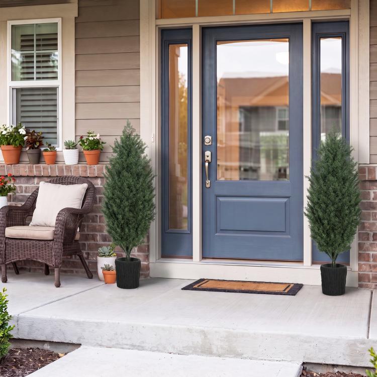 Two faux cedar trees with green branches in black textured pots at an entryway