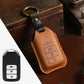 Tan leather key fob cover with black strap, metal ring loop, and embossed control buttons.