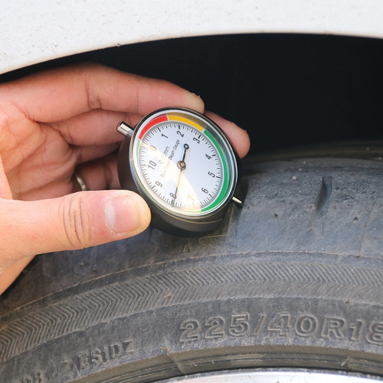 Hand holding tire tread depth gauge on car tire tread; pointer dial is visible