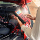 Hand holding red-and-black 12V battery tester connected to car battery terminals