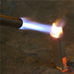Gas blow torch with blue flame and orange tip aimed at a metal surface