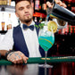 Stainless cocktail shaker pouring into a glass with green mixer at a bar