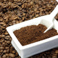 White container filled with ground coffee beside a manual stainless grinder