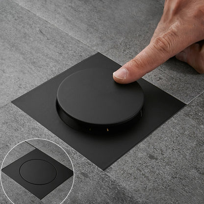 Foot-operated floor drain with black round lid on square base, pressed by hand