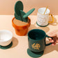 Cactus coaster next to cups: green felt cactus on cork base in an orange pot.