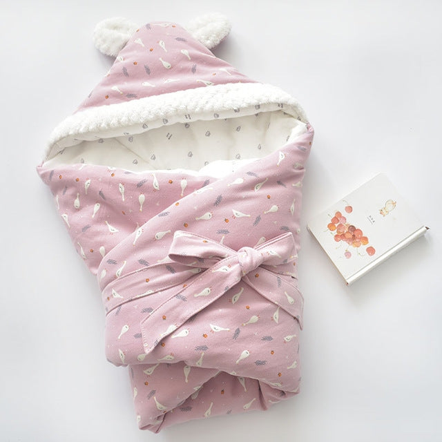 Baby holding blanket with pink cartoon print, white fleece trim, and tied bow hood