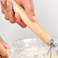 Hand holding an oak-handled coil whisk over a bowl of flour and dough