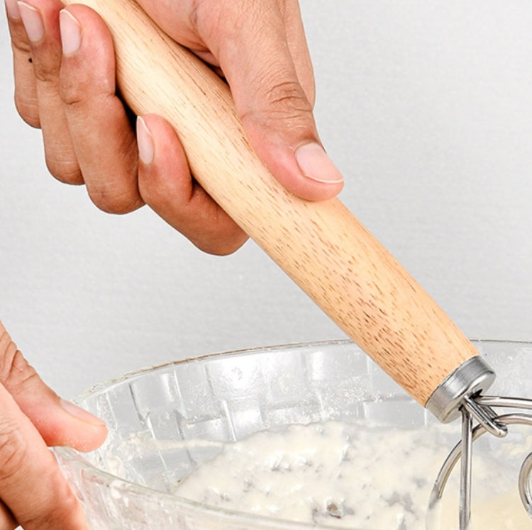 Hand holding an oak-handled coil whisk over a bowl of flour and dough