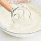 Oak-handled coil whisk mixing pale dough in a glass bowl