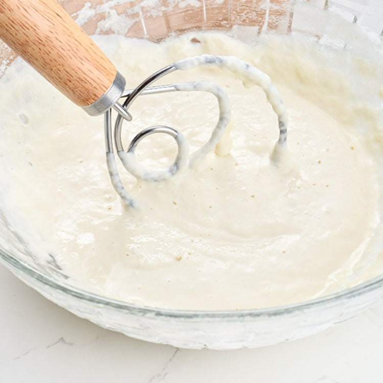 Oak-handled coil whisk mixing pale dough in a glass bowl