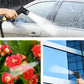 Hand using black sprinkler nozzle to spray water on cars, flowers, and building