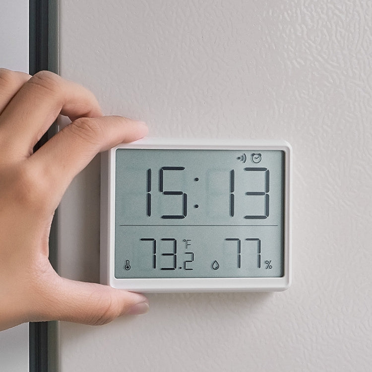 White LCD digital clock being mounted on a wall by hand; time and humidity shown