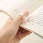 Hand holding a transparent acrylic thumb bookmark on the edge of an open book.