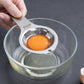 Hand holds stainless steel egg yolk separator over glass bowl with yolk inside