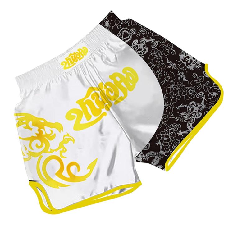 Muay Thai boxing shorts in white with black floral panel and gold swirl “2DROP” print.