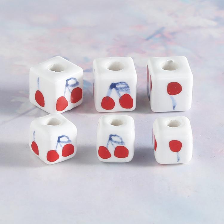 Square ceramic beads with red cherry and blue bow artwork, visible large center hole.