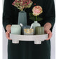 Hands holding round storage tray with wide rim, white base feet, decorative jars