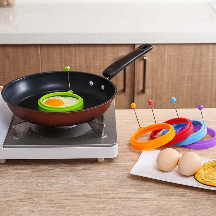 Green silicone egg ring mold in pan with assorted colored ring molds on table.